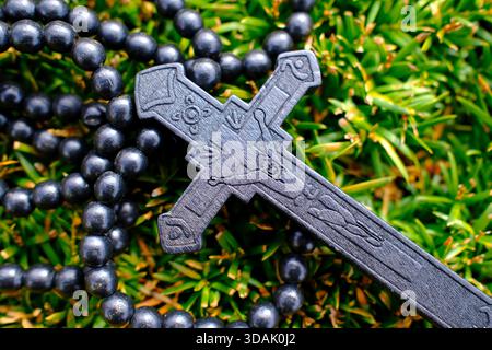 Dunkler Ebenholz-Rosenkranz mit aufwändig geschnitztem Kruzifix, das über blühendem gelbem Heidekraut auf üppig grünem Sträucher hängt und an friedliche christliche Hingabe erinnert Stockfoto