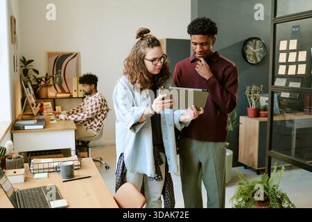 Junge kaukasische Frau zeigt dem jungen Schwarzen im modernen Büro Tablet Stockfoto