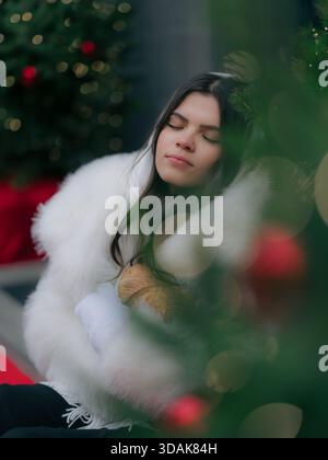 Nahaufnahme Porträt friedliche junge Frau mit geschlossenen Augen, mit kuscheliger weißer Winterjacke und Fäustlingen, gebackene Brötchen in der Nähe des festlichen Weihnachtsbaums. B Stockfoto
