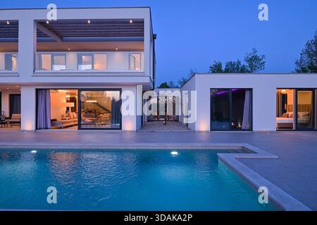 Blick von außen auf ein modernes Luxushaus mit Swimmingpool in der Dämmerung. Beleuchtete zweistöckige weiße Villa mit großen Glasfenstern, Terrasse und Terrasse Stockfoto