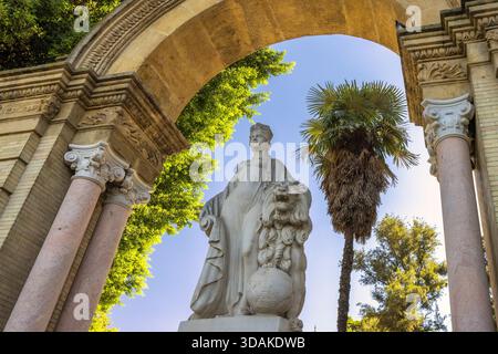 Sevilla, Spanien - 8. September 2024: Klassische Statue, eingerahmt von einem Bogengang, umgeben von üppigem Grün unter einem hellblauen Himmel. Stockfoto