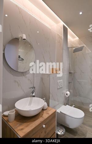 Modernes, elegantes Badezimmer mit Fliesen in weißer Marmoroptik, einem Holzwaschtisch mit Waschbecken, einer wandmontierten Toilette und einer beleuchteten Glasdusche Stockfoto