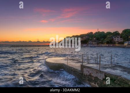 Wunderschöner Blick auf den Sonnenuntergang auf die Küste von Castiglioncello, Toskana, Italien, mit einem Betonpier oder einer Plattform, die ins Wasser hinausreicht. Wellen krachen um t herum Stockfoto