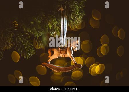 Schaukelnde Pferdedekoration, die einen Tannenzweig verziert, schafft eine herzerwärmende und nostalgische Urlaubsatmosphäre mit warmen Bokeh-Lichtern im Hintergrund Stockfoto
