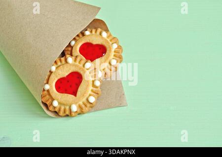 Shortbread-Kekse mit rotem Herzgelee in der gerollten Vintage-Papiertüte auf dem hellgrünen Holztisch. Nahaufnahme von Shortbread Keksen Stockfoto