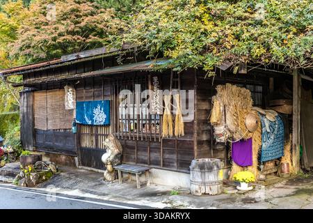 Ein kleines Geschäft mit Norenvorhang, einer großen Strohfigur und Herbstbäumen in Tsumago-juku, Nagiso, Präfektur Nagano, Japan. Strohdekorationen sind Teil der traditionellen Volkskunst der Region Kiso Valley Stockfoto