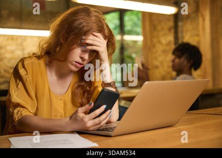 Rothaarige Frau, die den Kopf hält und auf das Telefon starrt Stockfoto
