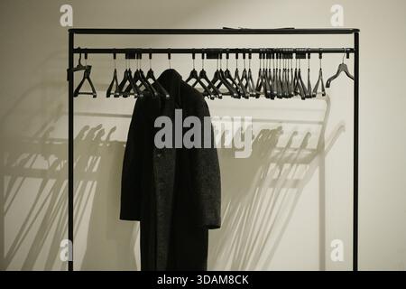 Ein langer schwarzer Mantel hängt an einem Kleiderbügel an einem mobilen Rack an einer weißen Wand Stockfoto