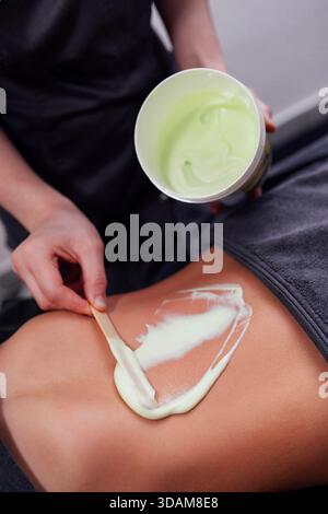 Der Spa-Therapeut trägt sanft grüne Creme mit einem Spatel auf den Rücken des Kunden auf und betont Entspannung und Hautpflege in einer ruhigen Wellness-Umgebung || Model veröffentlicht Stockfoto
