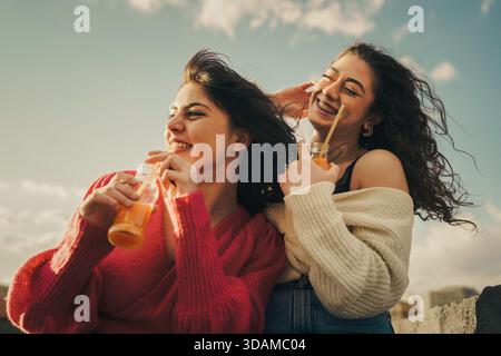 Zwei junge Freundinnen genießen einen glücklichen Moment zusammen im Freien, lachen und trinken erfrischenden Orangensaft. Stockfoto
