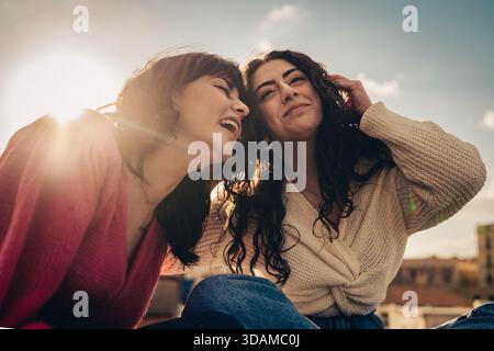 Zwei fröhliche junge Frauen lachen im Freien während einer schönen goldenen Stunde, beleuchtet von warmem Sonnenlicht und einem weichen Lichtstrahl, und feiern Stockfoto