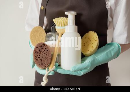 Person mit Handschuhen und Schürze, die natürliche Schwämme, Bambusbürste und Seifenflaschen zur Reinigung hält Stockfoto