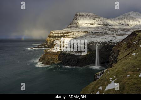 Der Blick auf schneebedeckte Klippen trifft auf das aufgewühlte Meer, ein Wasserfall taucht aus schneebedeckten Höhen unter einem wilden Himmel, Andeutungen eines Regenbogens prägen die Szene, Sor Stockfoto
