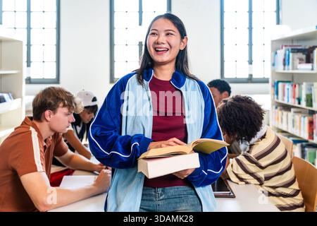 Junge Schülerin lächelt und lacht, während sie Bücher hält und selbstbewusst in ihren verschiedenen Klassenkameraden steht, die in einem Bright studieren und recherchieren Stockfoto