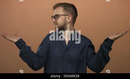 Mann mit Brille und einem marineblauen Hemd, der Handzucken im Studio gegen Pfirsichwand zeigt; Unsicherheit. Stockfoto