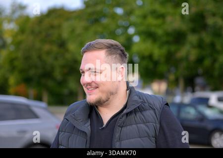 Porträt eines lächelnden jungen Mannes, der draußen eine schwarze Jacke trägt, an einem Herbsttag mit grünen Bäumen im Hintergrund. Stockfoto