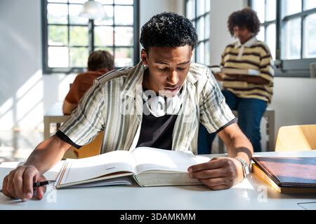 Der junge Mann konzentrierte sich auf das Studium eines Lehrbuchs an einem hellen Bibliothekstisch, konzentrierte sich auf Lehrveranstaltungen und Forschung mit Klassenkameraden, die im Hintergrund verschwommen waren Stockfoto