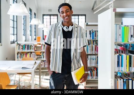 Ein junger schwarzer Student mit Kopfhörern um den Hals steht in einer hellen Universitätsbibliothek mit einem Buch und lächelnd, verkörpert das Lernen, vertraut an Stockfoto