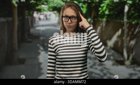 Frau mit Brille und gestreiftem Hemd zeigt mit dem Finger auf den Tempel auf der sonnendurchfluteten Straße mit schattigen Schatten; nachdenkliche Konzentration. Stockfoto