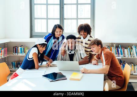Multiethnische Gruppe junger Studenten, die sich in der Bibliothek treffen - multiethnische Gruppe glücklicher junger Studenten, die gemeinsam an einem Laptop und Büchern in einem br Stockfoto