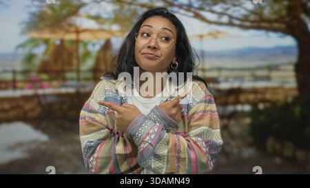 Frau mit gekreuzten Armen und Fingern, die in beide Richtungen auf Straßengeländer unter Bäumen zeigen, Brille und gemusterte Jacke tragen; Ungewissheit. Stockfoto