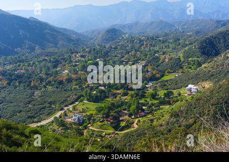 Der weitläufige Blick aus der Vogelperspektive fängt eine lebendige Wohnsiedlung ein, eingebettet zwischen üppigen grünen Bäumen und gewundenen Straßen in einem weitläufigen Bergtal. Stockfoto