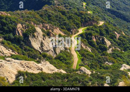 Serpentine Dirt Trail schlängelt sich durch zerklüftete, grüne Hügel und felsige Ausläufer im Red Rock Canyon Park, Santa Monica Mountains, unter einem klaren, hellen Licht Stockfoto