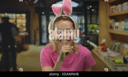 Frau in rosa Hemd mit Hasenohren lächelt und hält den Finger an die Lippen, um im Gebäude Ruhe zu finden; ruhige Geheimhaltung. Stockfoto