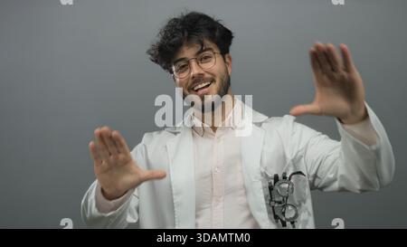 Junger hispanischer Mann mit Bart in Brille Lächeln und Gesten in einem weißen Mantel vor grauem Hintergrund, der Selbstvertrauen und Freundlichkeit zeigt. Stockfoto