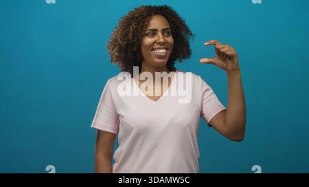 Junge afroamerikanische Frau, die im Studio mit türkisfarbenem Hintergrund eine kleine Lücke zwischen den Fingern einklebt; kleine Freude. Stockfoto