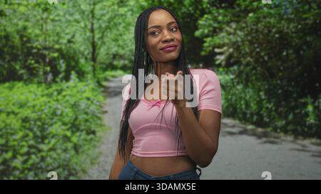 Die lächelnde Frau zeigt den Zeigefinger auf dem Waldweg mit rosa Crop Top und Jeans nach oben; Selbstbewusstsein stärken. Stockfoto