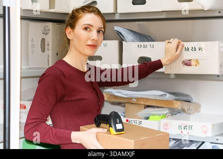 Die Frau in Burgunderbekleidung hält Box und Scanner, die Finger berühren den weiß gekennzeichneten Behälter auf einem hohen Regal in verpackungsgefülltem Lagerraum. Stockfoto