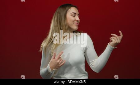 Die junge blonde Frau imitiert spielerisch das Gitarrenspiel vor einem leuchtend roten Hintergrund und zeigt einen lässigen und fröhlichen Ausdruck in einer isolierten Umgebung. Stockfoto