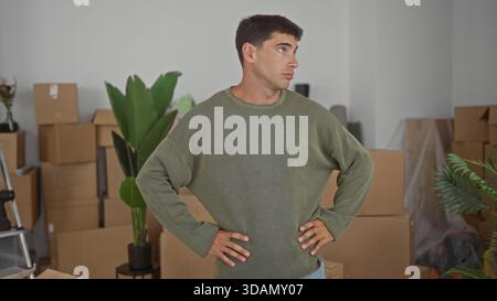 Junger Mann steht mit Händen auf Hüften neben gestapelten Pappschachteln im Innenraum des Gebäudes und trägt einen olivfarbenen Pullover; Unsicherheit. Stockfoto