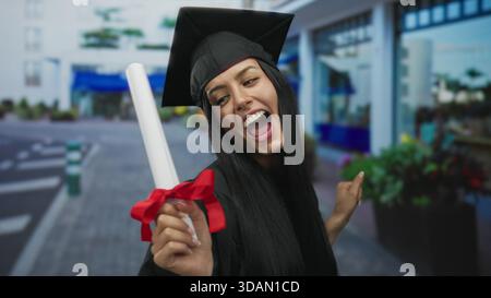 Frau, die den Abschluss im Freien in der Stadtstraße mit Diplom und Abschlusskappe feiert, zeigt junge hispanische Frau in akademischer Kleidung lächelnde Freude Stockfoto