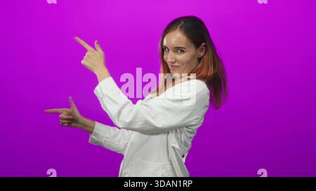 Frau Ärztin in weißem Mantel zeigt Finger wie Fingerpistolen im Studio, zeigt Hände und Zeigefinger zeigende Geste; spielerisches Selbstvertrauen. Stockfoto