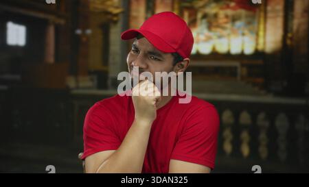 Mann in roter Mütze und Hemd steht drinnen mit Hand am Kinn im Kirchengebäude, Augen knirschen, während Buntglas die Bänke beleuchtet; Sorge. Stockfoto