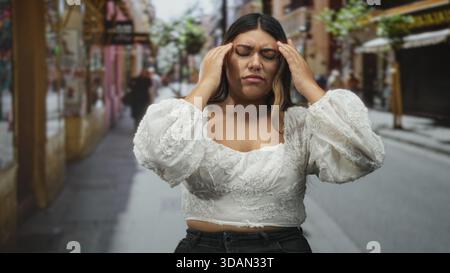 Frau mit Hand auf der Stirn für Kopfschmerzen auf der Straße, mit weißer, bestickter Bluse, zuwinkenden und reibenden Schläfen; Stress-Müdigkeit-Frustration. Stockfoto