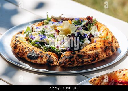 Gourmet-Pizza mit einer dicken, flauschigen Kruste, dekoriert mit essbaren Blumen und frischem Gemüse, serviert im Freien bei natürlichem Sonnenlicht. Stockfoto