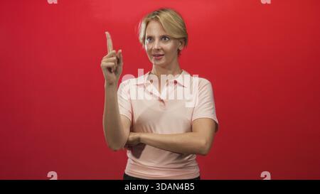 Frau zeigt den Finger nach oben mit gefaltetem Arm in der roten Wand des Studios, Zeigefinger als Geste hochgehoben; Selbstbewusstsein. Stockfoto