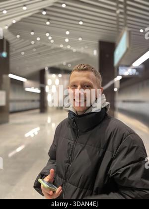Ein lächelnder junger Mann und ein Smartphone in der Hand, während er in einer hellen, modernen U-Bahn-Station steht. Stockfoto