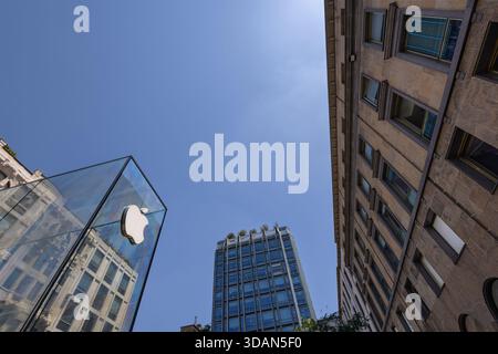 MAILAND, ITALIEN, 06.19.2025: Ein moderner Apple Store verfügt über eine hohe Glasfassade neben einem historischen Gebäude vor einem klaren blauen Himmel. Die Szene verschmilzt Stockfoto