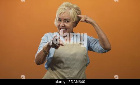 Die ranghöchste hispanische Frau in der Schürze zeigt den Finger auf die Kamera und zeigt den Finger zum Tempel in einem Studio mit orangefarbenem Hintergrund; spielerisches Selbstvertrauen. Stockfoto