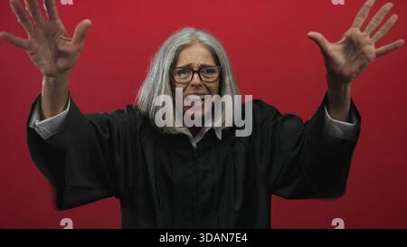 Grauhaarige Richterin mit schwarzem Gewand und Brille hebt die Hände, um das Gesicht im roten Studio zu schützen; Frustration. Stockfoto