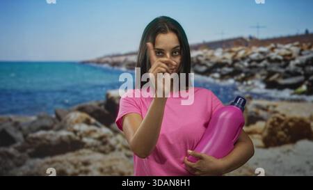 Junge Frau, die mit dem Zeigefinger auf die felsige Küste zeigt und eine große pinkfarbene Waschmittelflasche hält; verspielte Putzbotschaft. Stockfoto