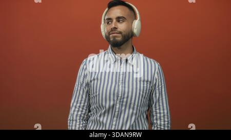 Junger hispanischer Mann mit weißen Kopfhörern und gestreiftem Hemd im Studio vor der orangen Wand; verspielt. Stockfoto
