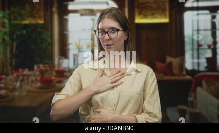 Frau mit Brille und beigefarbener Bluse berührt die Brust im Gebäude mit ruhiger Geste, während sie sich auf ihren Atem konzentriert; Gelassenheit. Stockfoto