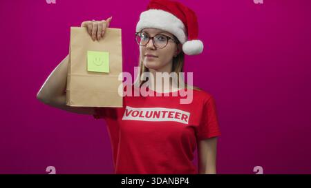 Freiwillige Frau mit weihnachtsmütze zeigt mit dem Finger auf die Papiertüte mit Smiley-Note in einem rosa Studio; Großzügigkeit. Stockfoto