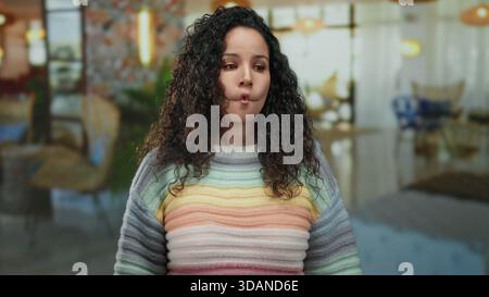Hispanische Frau mit lockigem Haar, die drinnen in einem Hotel steht und einen farbenfrohen gestreiften Pullover trägt, der verschiedene nachdenkliche Posen gegen einen bl ausdrückt Stockfoto