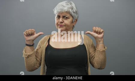 Frau zeigt in einem grauen Studio die Daumen auf die Brust, trägt Tanktop und Strickjacke mit Basketball und Armbändern; Selbstvertrauen. Stockfoto